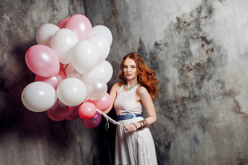 Red-haired beauty young woman in a white dress, holding a large bundle of balloons at the party.