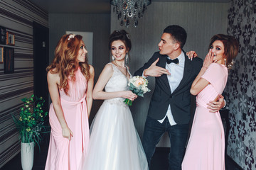 Cheerful young newlyweds along with cute bridesmaids. Stylish wedding at home. Young cute newlyweds hug along with bridesmaids at home