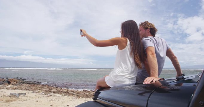 Couple taking selfie using smartphone at Shipwreck beach. Male and female hikers are sitting on offroad vehicle car trunk enjoying summer vacation on Lanai, Hawaii, USA. RED EPIC SLOW MOTION.