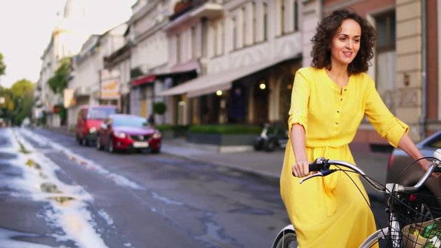 Beautiful woman riding a city bicycle with a basket and flowers in the city center while talking to someone. Steadicam shot. Beautiful old city view