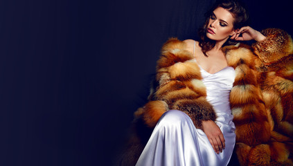 A gorgeous girl with long brown hair and a white satin dress-combination sits in a fur coat. The...