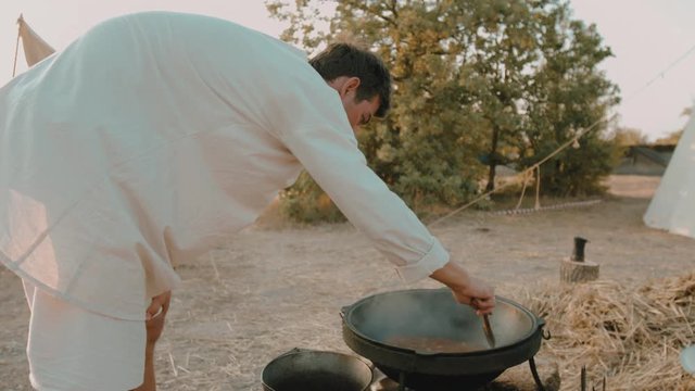 Medieval man stirs the pottage with meat in the cauldron on the fire