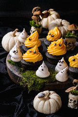 Halloween decorated sweet dessert table black cupcake with orange cream, white meringue ghosts with chocolate eyes, decor skulls and pumpkin on clay plate  over black background.
