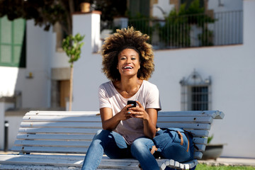 Fototapeta premium beautiful young african woman sitting outdoors on bench with mobile phone