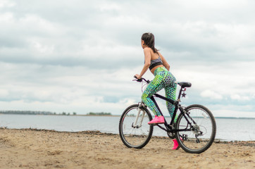 Obraz premium Girl beautiful brunette. Athletic body , pumped muscles. Bright colors , green ,pink . riding her bike along the shore. On the dirty sand