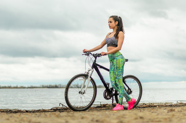 Fototapeta premium The girl the brunette plays sports on the seashore - on the beach. Rides a bike.