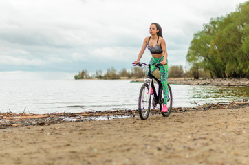 Fototapeta premium The girl the brunette plays sports on the seashore - on the beach. Rides a bike.