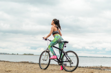 Fototapeta premium The girl the brunette plays sports on the seashore - on the beach. Rides a bike.