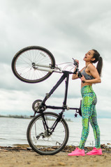 The girl the brunette plays sports on the seashore - on the beach. Rides a bike.