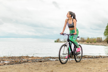 The girl the brunette plays sports on the seashore - on the beach. Rides a bike.