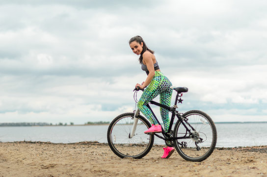 The girl the brunette plays sports on the seashore - on the beach. Rides a bike.