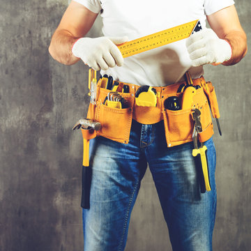 unidentified handyman standing with a tool belt with construction tools and holding roulette against grey background, toned image. DIY tools and manual work concept