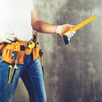 unidentified handyman standing with a tool belt with construction tools and holding roulette against grey background, toned image. DIY tools and manual work concept