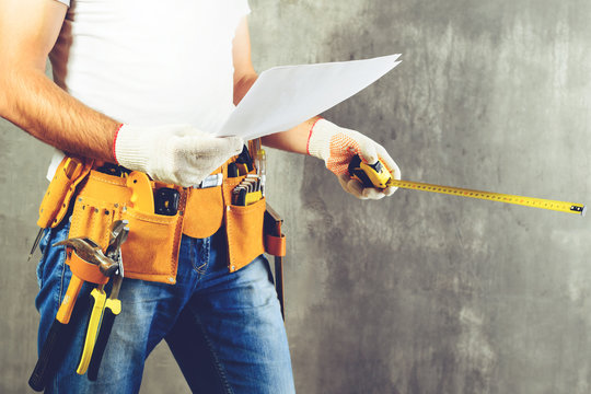 unidentified builder standing in white gloves with a tool belt with construction tools and holding roulette and the project plane against grey background, toned image