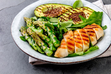 Healthy grilled chicken, grilled avocado and asparagus salad with linen seeds. Balanced lunch in bowl. Gray slate background. Top view