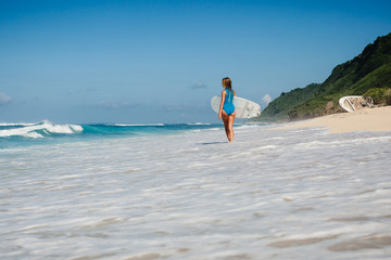 Female surfer in blue swimwear with board in hands on coastline