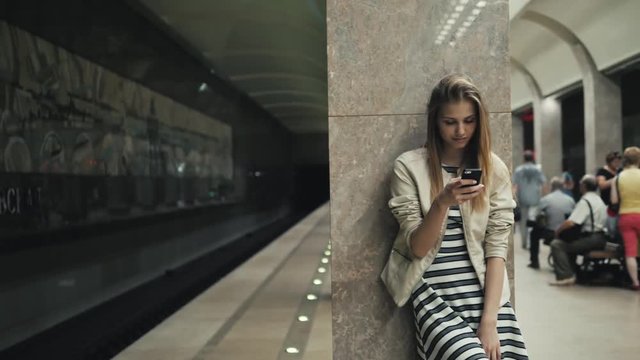 Girl with smart phone at train station. Portrait of a young caucasian woman, looking at the phone while waiting for the train on the platform, subway transportation public