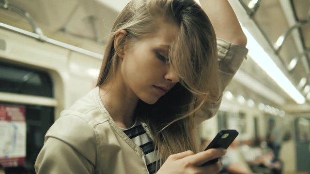Portrait of a smiling lovely girl typing message on mobile phone in subway train, sexy student blonde woman with smartphone happy smiling