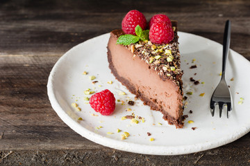 Slice of chocolate cheesecake decorated with pistachios, raspberries and mint leaf on white plate on wooden background.