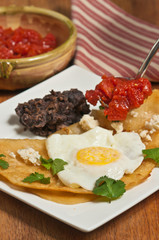 Huevos rancheros, meal, fried taco, fried egg,  chopped tomatoes, refried beans, parsley on a square, white plate, bowl of tomatoes, a red and white stripped napkin, wood serving board