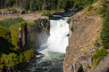Lower Mesa Falls in the Caribou-targhee National Forest with mist being splashed onto the green foliage and rock tower.