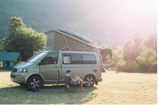 Nomad adventurer with pet dog rest on camping site after long day of driving, sit on folding chair looks at mountains and sunset, next to camper van with bike rack