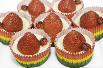 Cheesecake cupcakes in red, yellow, and green paper cups topped with red strawberries and chocolate candy ladybugs on a white plate.
