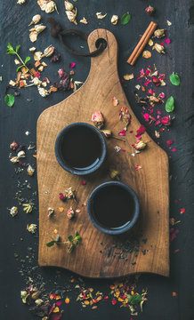 Chinese black tea in black stoneware cups on serving wooden board over black wooden background with herbs, flower buds, tea leaves spilt around, top view
