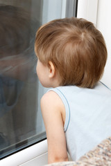 A little boy with blond hair in his home clothes looks in a dark window