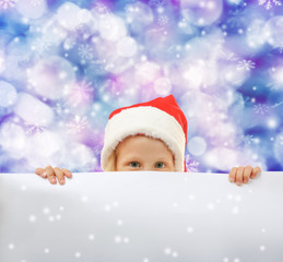 Cute little girl in Santa hat hiding behind blank poster on blurred lights background