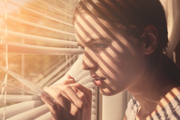 Beautiful young girl separating slats of blinds and looking through window
