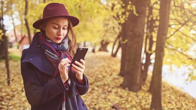 Young Woman Using SmartPhone in Autumn Park. SLOW MOTION. Girl dressed in wool coat and hat enjoying cool fall weather outdoors, texting and taking photos on smart phone. Golden falling leaves
