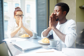Fototapeta na wymiar Multi-ethnic couple gathered around table and praying before having breakfast, homemade croissants and fragrant coffee waiting for them