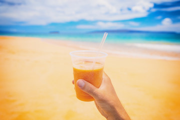 Healthy juice smoothie juice drink selfie on beach. Turmeric, carrot, papaya or orange fruit, Man hand holding plastic cup on beach summer background. Food picture of tropical fruits diet lifestyle.