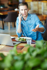 Young man speaking on smartphone by lunch in cafe with laptop in front