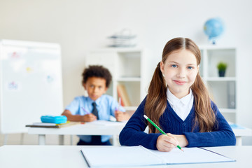 Adorable schoolgirl with crayon sitting by desk with classmate on background