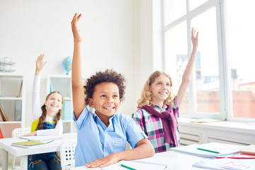 Happy and clever learners of elementary school raising their hands at lesson