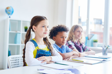 Diligent girl and her classmates listening to teacher explanations by desk