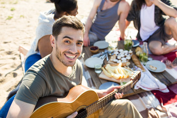 Smiling guy with guitar playing it with his friends on background