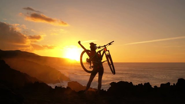 Aspirations, success and achievement. Active lifestyle MTB cyclist mountain biking woman cheering happy raising arms lifting bike by sea at sunset celebrating. Person with bicycle in amazing nature.