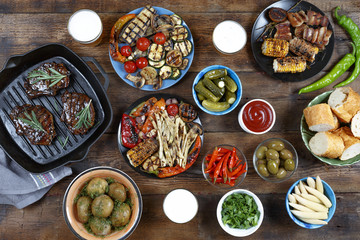 Dinner table with meat grill, bbq vegetables, salads, sauces, snacks and beer, top view