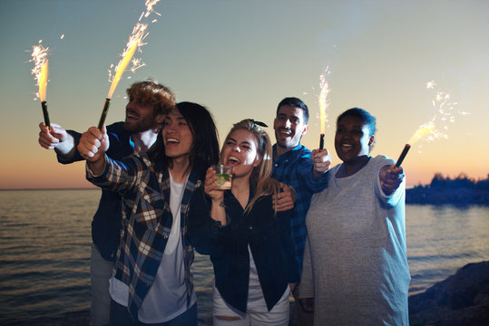 Group of ecstatic friends with fire-crackers having fun by seaside in summer night
