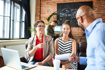 Group of cheerful managers analyzing results of accomplished work while having working meeting, interior of modern open plan office on background