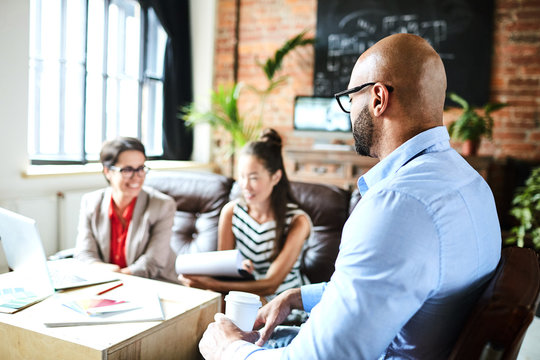 Profile view of bearded mixed race man with paper cup of coffee in hands sitting on cozy armchair and discussing ambitious project with colleagues, interior of modern open plan office on background