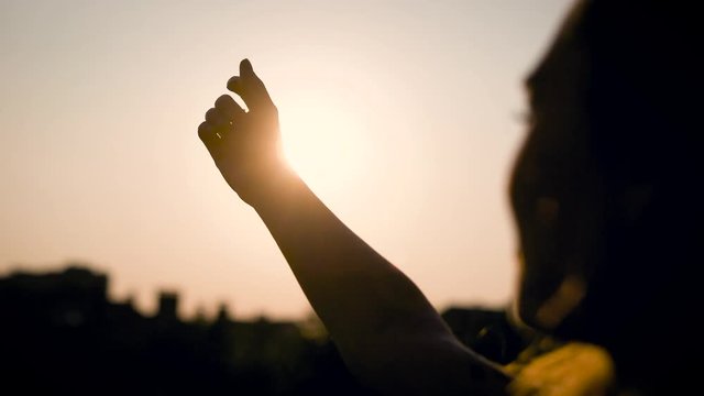 thoughtful young woman is trying to close the sun by hand in summer evening, silhouette of face and hand