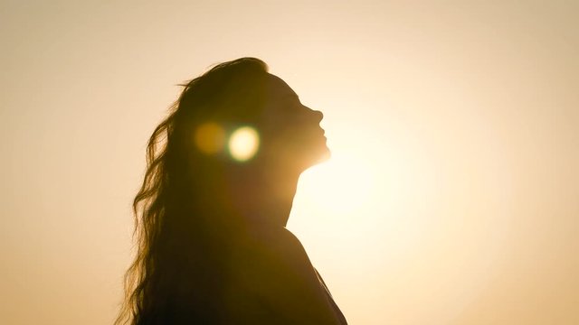 young woman with long wavy hair is wearing in summer dress is standing against the sun in evening time