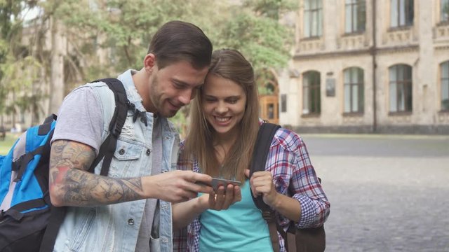 Cute young students watching images on smartphone on campus. Handsome brunette man holding cellphone in his hands. Pretty caucasian brown haired girl gesturing with her hands