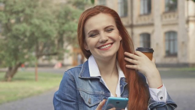Pretty readhead female student laughing at what she seeing on her smartphone on campus. Close up of attractive young woman holding cup of coffee and cellphone in her hands. Cute caucasian girl