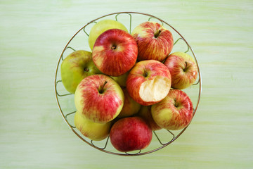 Metal bowl with green, yellow and red apples  and one bitten apple on the green wooden background, top view.