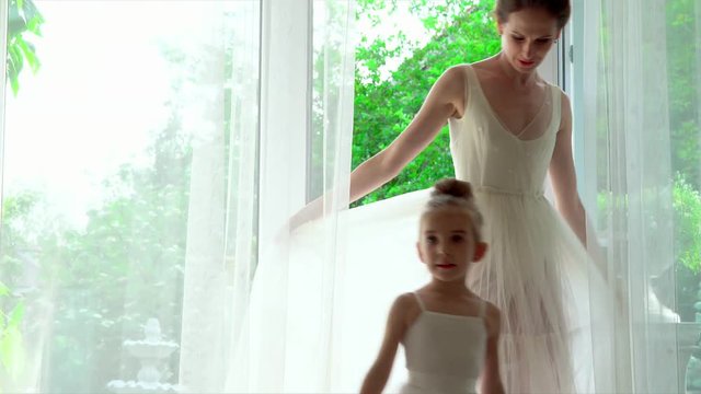The little ballerina will repeat after the teacher. Two ballerinas are by the big window with white curtains. Beautiful picture. The teacher teaches the little ward..